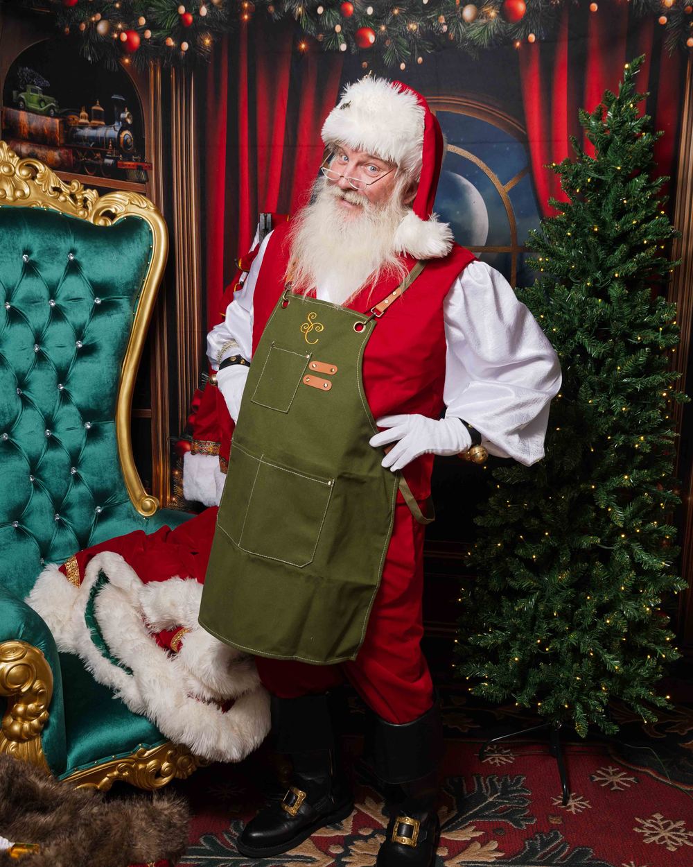 Santa Doug in his red suit and green workshop apron, standing between a gold throne and a lit Christmas tree