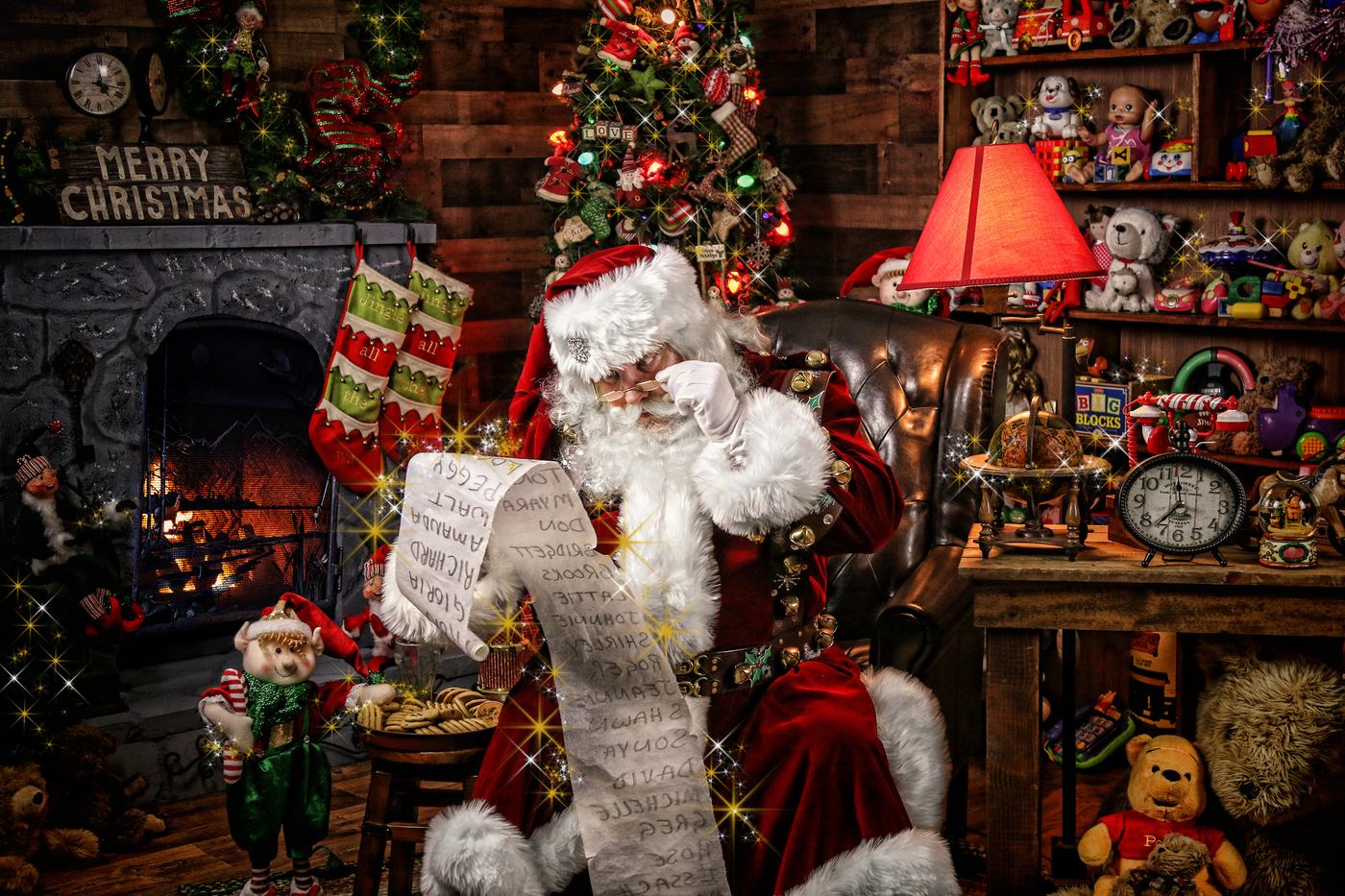 Santa Doug checking his long list in his workshop, stockings and a lit tree behind him